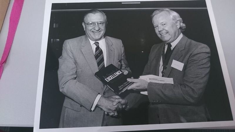 Archive photograph of two men shaking hands while presenting a copy of the book In Search of Excellence, with Institute of Administrative Management papers visible